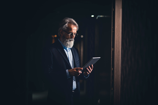 Senior businessman standing in a dark hotel room and using a digital tablet