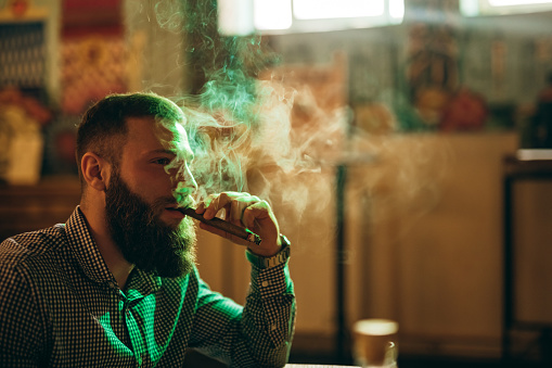 Portrait of Handsome bearded Caucasian elegant man smoking cigar in bar.