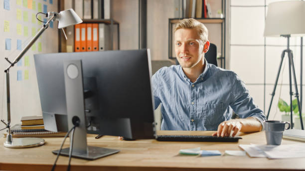 istock Creative Entrepreneur Sitting at His Desk Works on Desktop Computer in the Stylish Office. Handsome Caucasian Man Uses Computer. In the Background Stylish Office Studio with Modern Minimalism Design 1268697895