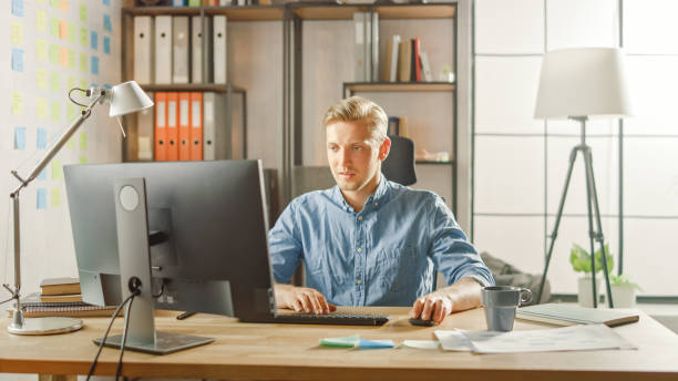 istock Creative Entrepreneur Sitting at His Desk Works on Desktop Computer in the Stylish Office. Handsome Young Businessman Uses Computer, Does Outsourcing Job, Designs New Apps and Develops Software 1268697897