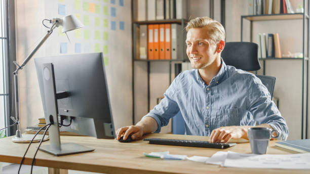 istock Creative Entrepreneur Sitting at His Desk Works on Desktop Computer in the Stylish Office. Handsome Caucasian Man Uses Computer. In the Background Stylish Office Studio with Modern Minimalism Design 1268697913