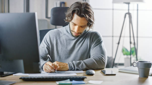 istock Handsome Long Haired Entrepreneur Sitting at His Desk in the Office Works on Desktop Computer, Working with Documents, Charts, Graphs, Statistic and Company Strategy. Businessman Fills Documents 1268697915