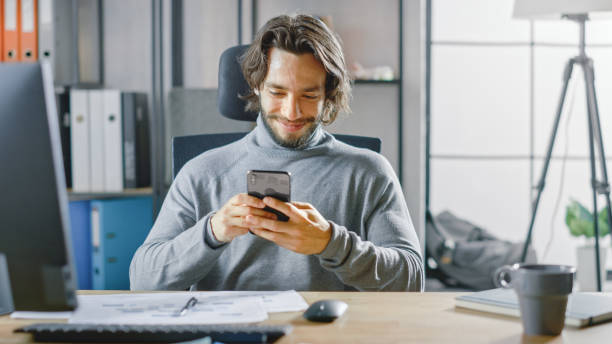 istock Handsome Long Haired Entrepreneur Sitting at His Desk in the Office Works on Desktop Computer, Working with Documents, Graphs. Uses Smartphone, Social Media App, Writing Emails, Messaging 1268697916