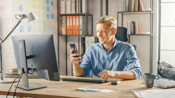 istock Creative Entrepreneur Sitting at His Desk Works on Desktop Computer in Stylish Office, Picks up and Starts Using Smartphone, Uses Social Media App, Emailing Business Partners, Messaging Family Members 1268697924