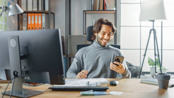 istock Handsome Long Haired Entrepreneur Sitting at His Desk in the Office Works on Desktop Computer, Working with Documents, Graphs. Smilingly Uses Smartphone, Social Media App, Writing Emails, Messaging 1268697927