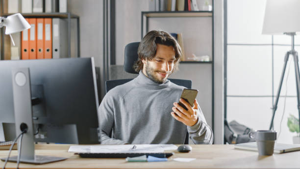 istock Handsome Long Haired Entrepreneur Sitting at His Desk in the Office Works on Desktop Computer, Working with Documents, Graphs. Uses Smartphone, Social Media App, Writing Emails, Messaging 1268697931