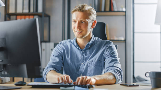 istock Handsome Blonde Entrepreneur Sitting at His Desk in the Office Works on Desktop Computer, Working with Documents, Charts, Graphs, Statistic and Strategy. Creative Businessman Using Computer 1268697943