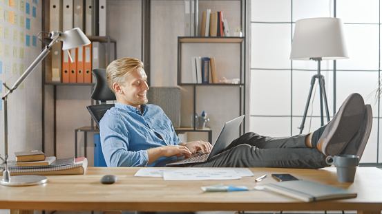 Handsome Young Entrepreneur Works on a Laptop with His Legs Up on the Desk. Designer Uses Notebook to Create Software Unicorn Startup. Chill Student Writing Paper for University