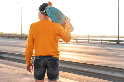 Stylish young man with a longboard on a bridge at sunset