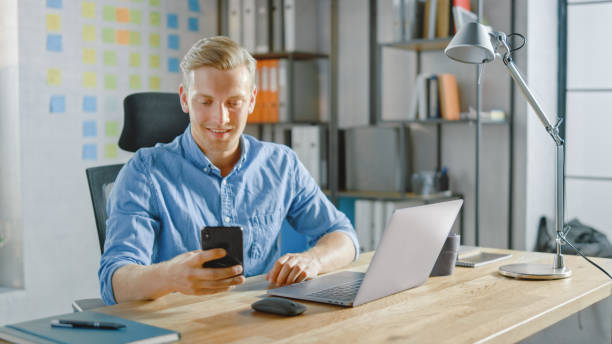 istock Creative Entrepreneur Sitting at His Desk Works on Desktop Computer in the Stylish Office, Picks up and Starts Using Smartphone, Uses Social Media App, Emailing Business Partners, Messaging Friends 1268703834