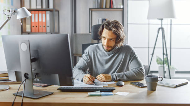 istock Handsome Long Haired Entrepreneur Sitting at His Desk in the Office Works on Desktop Computer, Working with Documents, Correcting Charts, Drawing Graphs, Statistic and Updating Company Strategy 1268703835