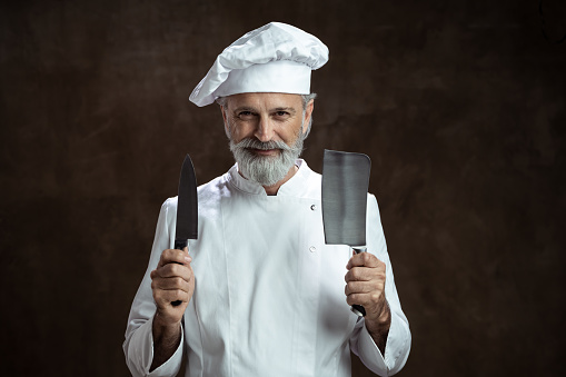 Adult man in a chef's whites, white hat and white double-breasted jacket holding knives on black background. Advertisement portrait for butchery or knives.