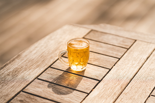 A glass of cold beer, filled to the top. Standing outdoors in the sun, on a wooden table.