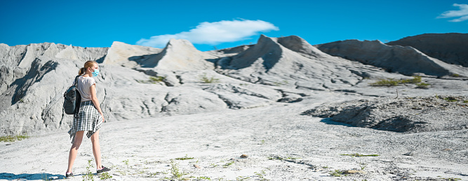 Young blonde woman walking against the white mountains wearing face protective mask for Covid 19 prevention. Young student traveler tourist with backpack. Corona virus and domestic tourism concept.