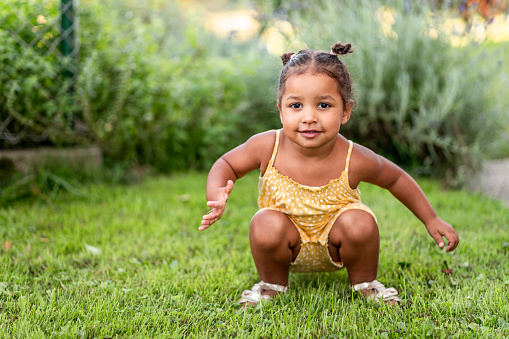 Portrait of a cute little mixed raced girl with two small hair buns on her head, wearing a yellow summer rummper, crouching on the green grass and looking at camera.