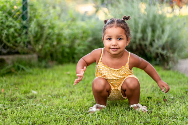 Little girl crouching on the grass Portrait of a cute little mixed raced girl with two small hair buns on her head, wearing a yellow summer rummper, crouching on the green grass and looking at camera. baby squatting stock pictures, royalty-free photos & images