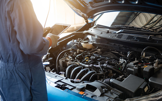 Mechanic in blue workwear uniform holding tablet and looking up with blur car in the background. Automobile repairing service, Professional occupation teamwork. Vehicle maintenance.