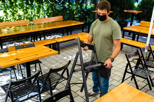 Diligent male cafeteria worker organizing tables and chairs at outdoor  coffee shop so everything can be by the rules of social distancing during COVID-19 pandemic