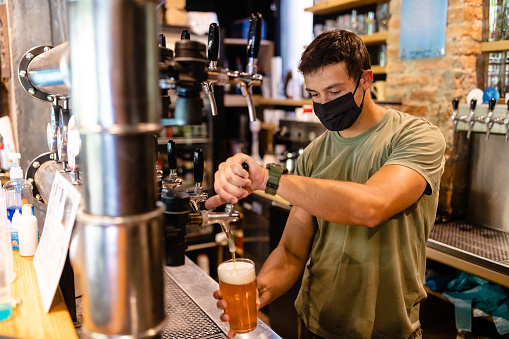 Waiter filling beer glass with ale from the beer tap inside the bar counter while wearing protective face mask during COVID-19 pandemic