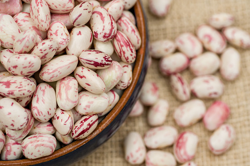 Close up of uncooked borlotti beans showing maroon marbling colour, in a blue ceramic earthenware bowl against a hessian background.  Borlotti beans are used in rustic Italian cooking, also known as cranberry beans.