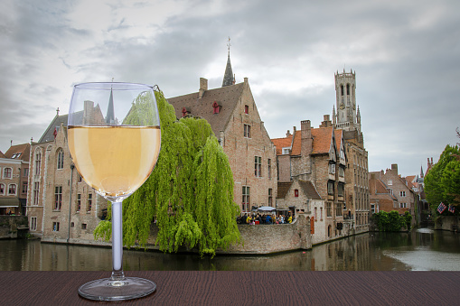 Glass of white wine with view from famous viewpoint in Bruges. View on Bruges old town and Belfry tower with pink sky during twilight, Bruges, Belgium.