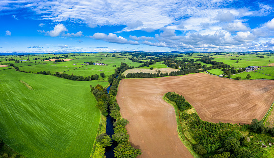 The view from a drone of a small Scottish river in a rural area of Dumfries and Galloway, south west Scotland, The river is in an area of farmland with fields on either side of it.\nThe panorama was created by merging several images together.