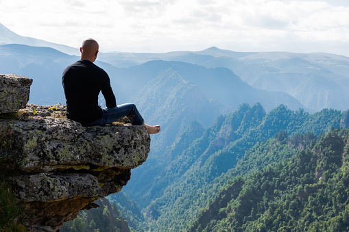 A young athletic man with a shaved head sits on top of a cliff and looks out at the horizon, enjoying the magnificent mountain range. Being on the edge of a cliff meditation in harmony with nature. Camp at an altitude of in the mountains.