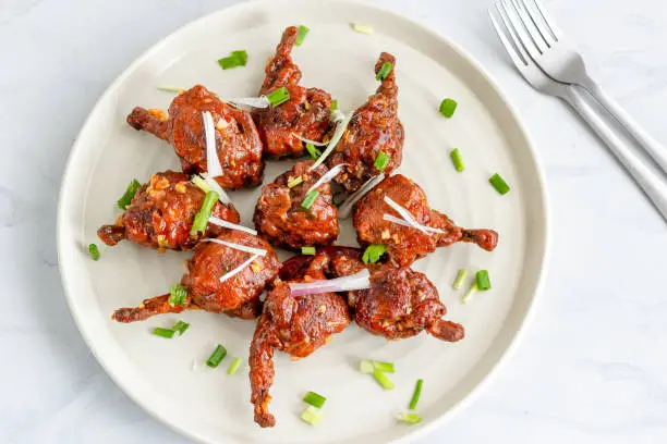 Popular Indo-Chinese Chicken Appetizer Made of Chicken Winglets on a White Plate on White Background Garnished with Scallion and Red Chili PepperHorizontal Top View Photo