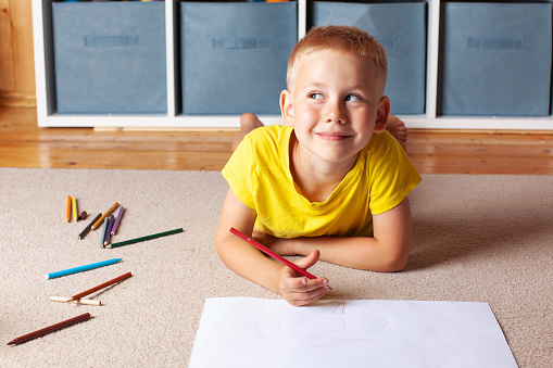 A blond boy in a yellow t-shirt lies on the floor with a drawing sheet and pencils and smiles thoughtfully.