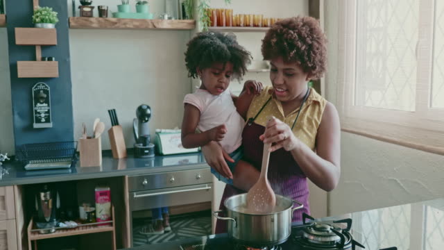 Real time video of Afro-Caribbean mother in mid 20s standing at stove holding preschool age daughter and showing her how to cook pasta.