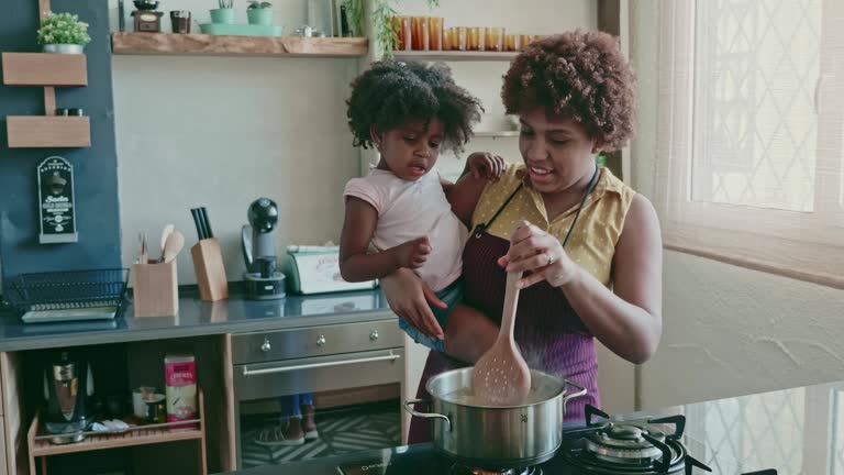 Real time video of Afro-Caribbean mother in mid 20s standing at stove holding preschool age daughter and showing her how to cook pasta.