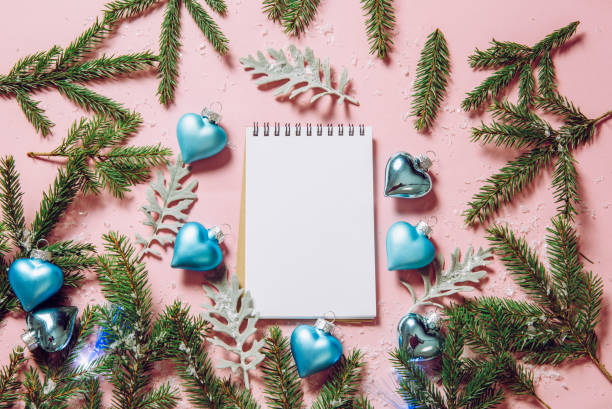 Flat lay view of empty notebook for check list with blue heart shape Christmas baubles and fir tree branches for decoration on light pink background, studio shot.