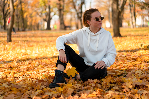 Woman in a white sweater with a hood sits on ground in the park and holding a leaf in her hands. Cute girl in white hoodie is sitting on the yellow leaves carpet