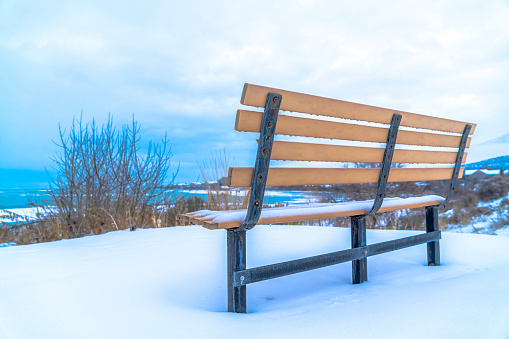 Frosted bench on snowy hill with Utah Lake and overcast sky view in winter. The empty bench has brown back rest and seat supported by metal frames.