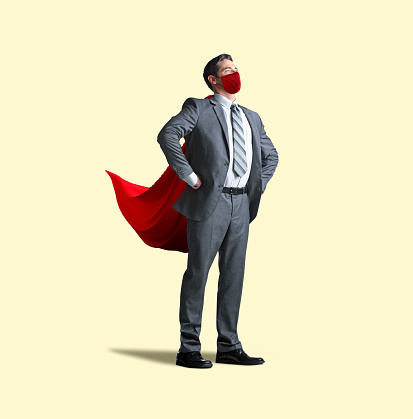 A man stands confidently with his hands on his hips as he wears both a red cape and a red protective face mask isolated on a yellow background.