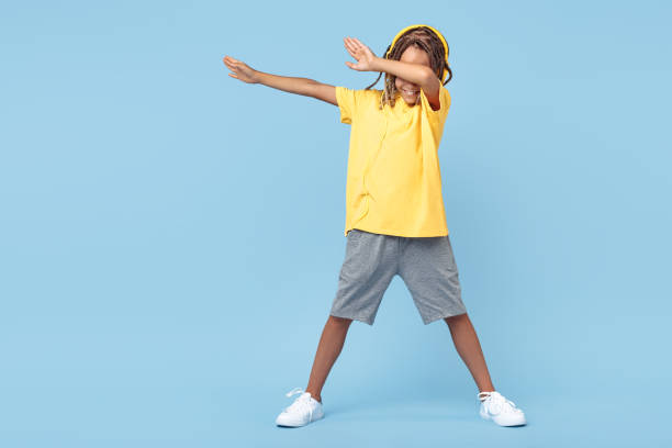 Cheerful little boy with african with headphones dabbing with one leg on soccer ball over blue background. Cheerful little boy with african with headphones dabbing with one leg on soccer ball over blue background dab dance stock pictures, royalty-free photos & images