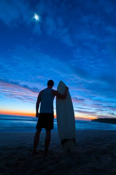 Silhouette of a man holding a surfboard at dawn. He is looking at the ocean waves with beautiful sunrise colours in the sky.