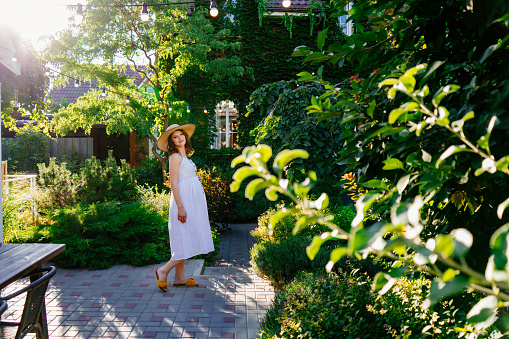 a pregnant woman in a white dress and hat walks in the back garden of a house. walking in the fresh air and a healthy lifestyle during pregnancy.
