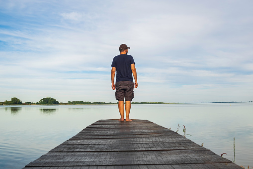 Man in short cargo pants standing on wooden pier and observing the surface of lake, Varazdin lake, Croatia