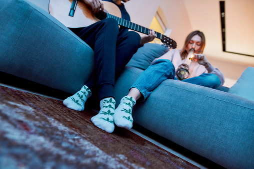 Young guy, artist writing song, playing guitar, sitting with his girfriend on the couch at home. Girl smoking marijuana from glass water pipe or bong. Weed legalization concept. Medical marijuana