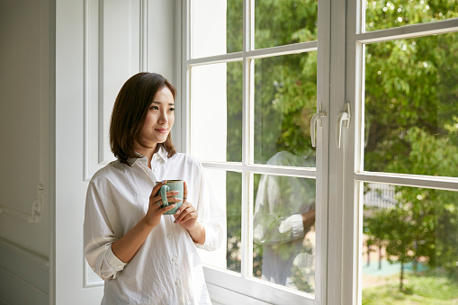 young asian woman standing by window at home holding a cup of coffee looking happy and content