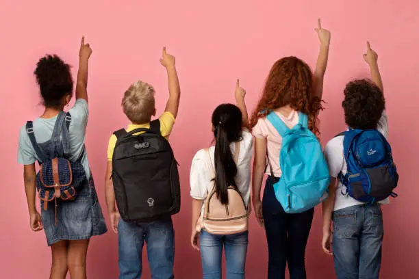 Back view of schoolkids with backpacks pointing up or touching virtual screen over pink background