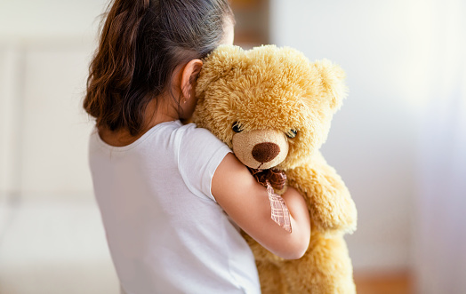 Unrecognizable Kid Girl Embracing Teddy Bear Toy Standing Alone Near Window Indoor. Loneliness Concept. Back View, Selective Focus