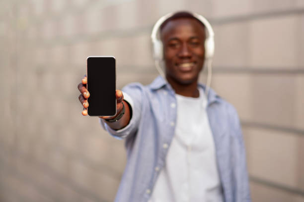 istock Millennial black man in headphones showing smartphone with empty screen outdoors, selective focus. Mockup for design 1269037395
