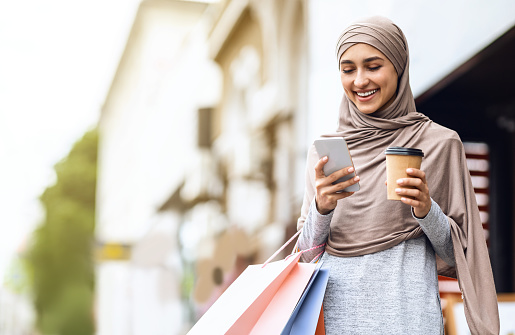 Cheerful arab girl in hijab with shopping bags and cup of coffee using mobile phone, spending weekend at shopping mall, empty space
