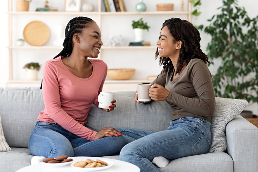 Black girlfriends enjoying weekend together, drinking coffee and talking on couch at home