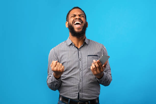 ragazzo nero eccitato che tiene il telefono e gesticola - shaking fist foto e immagini stock