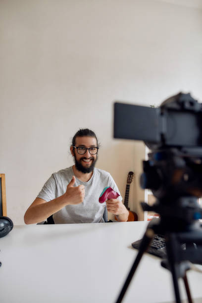 istock Male technology blogger in glasses showing thumbs up, holding game controller joystick while recording video blog or vlog about new gadgets at home 1269045561