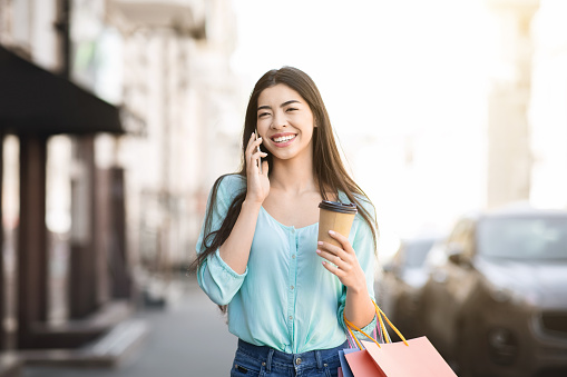 After Successful Shopping. Portraif Of Cheerful Asian Girl Walking With Shopper Bags And Coffee And Talking On Celphone Outdoors