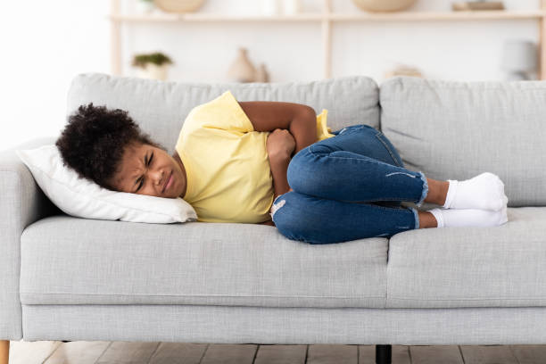 Black Girl Suffering From Abdominal Pain Lying On Sofa Indoor Stomachache. Black Teen Girl Suffering From Abdominal Pain Touching Aching Stomach Lying On Sofa At Home. stomachache stock pictures, royalty-free photos & images